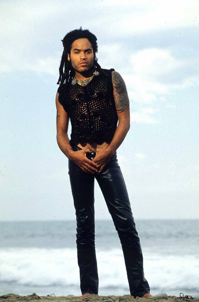 Lenny Kravitz standing on a beach, wearing a sleeveless crochet top and leather pants. He has long dreadlocks and tattoos on his right arm