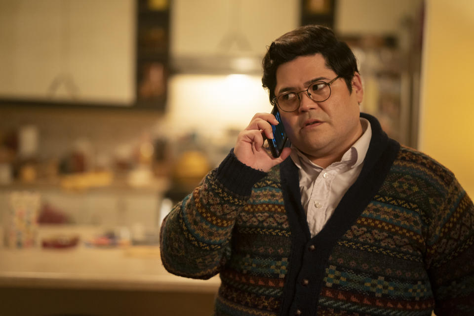 A man in a patterned button-down sweater and glasses, speaking into his cellphone in a kitchen; still from "What We Do in the Shadows"