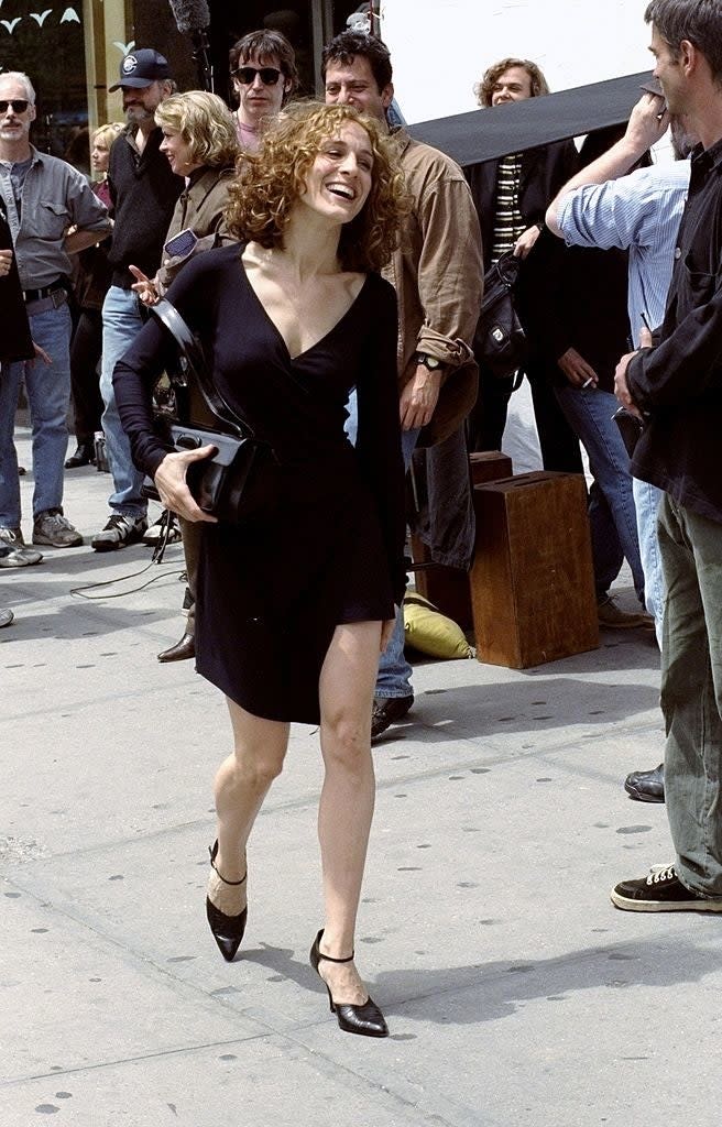 Sarah Jessica Parker walking on the sidewalk, smiling, in a black dress with long sleeves and black heels, surrounded by people