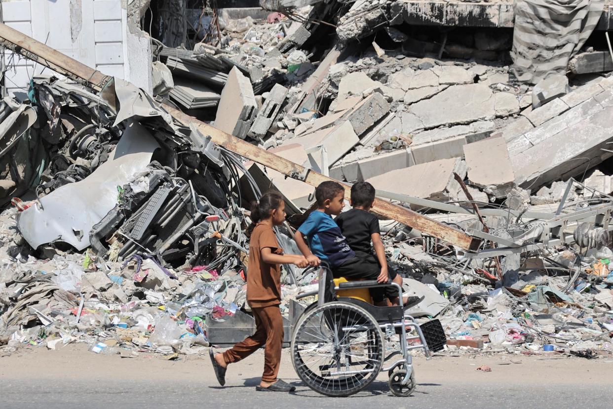 <span>A girl pushes a wheelchair with children sitting atop jerrycans past a collapsed building in Gaza City on 27 August 2024.</span><span>Photograph: Omar Al-Qattaa/AFP via Getty Images</span>