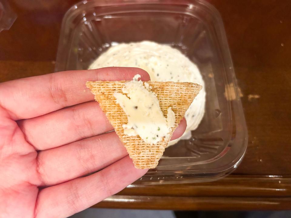 A hand holding a triangle cracker with a white butter with pieces of herbs in it in front of a container of butter