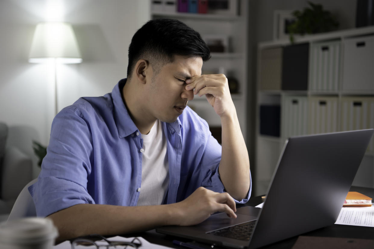 Young Asian man tired from hard work with eyes closed using hands to pinch and massage eyes and nose to relieve exhaustion and pain.