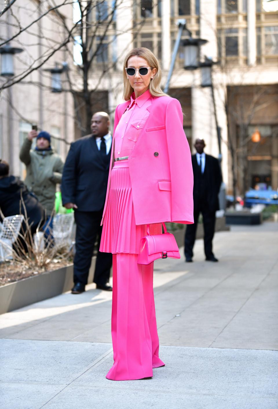 Celine in a bright pink monochrome ensemble. She's wearing a blazer over her shoulders, a button down top, and a pleated skirt over flared trousers. She's holding a pink purse and black sunglasses.