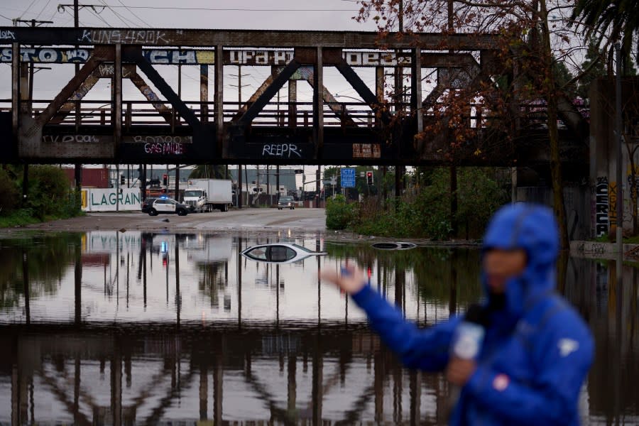 A television reporter does a broadcast as cars are seen submerged on a flooded street under a railroad bridge Thursday, Feb. 1, 2024 in Long Beach, Calif. (AP Photo/Eric Thayer)