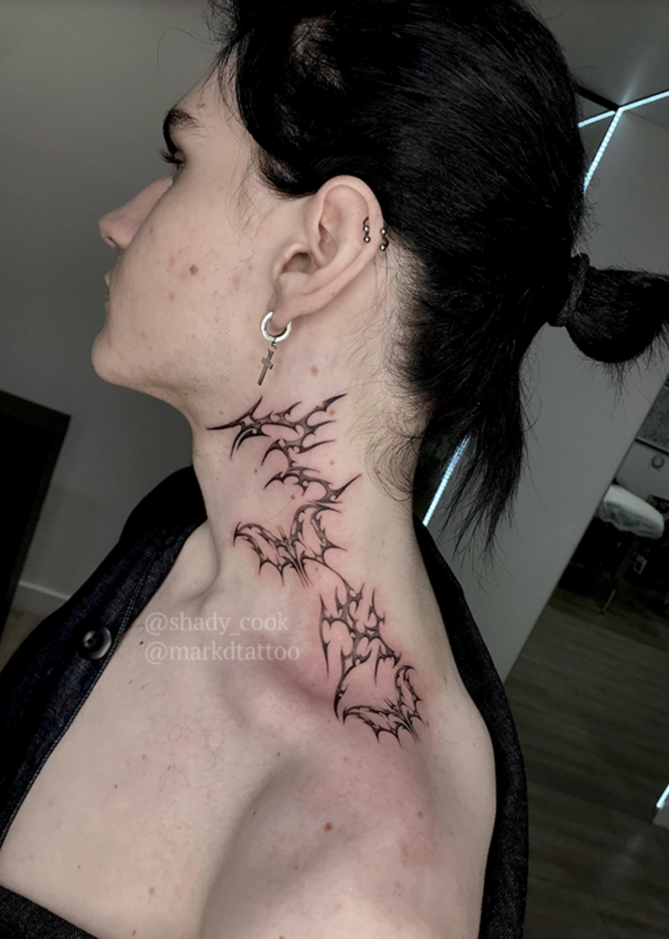 Side view of a person's neck with intricate tattoo art featuring jagged bat shapes from below the ear to the shoulder. The person's hair is tied back. Tattoos by @shady_cook and @markdtattoo