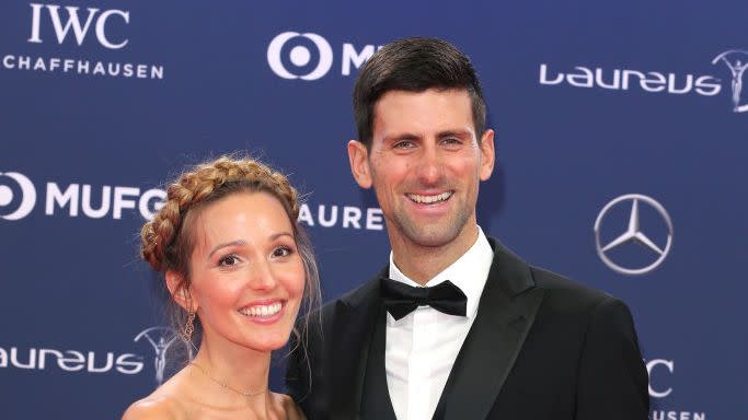 jelena and novak djokovic pose for a photo and smile at the camera, she wears a gray strapless stress and he wears a black tuxedo