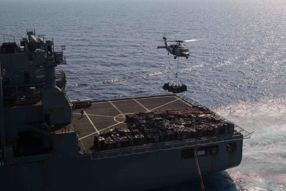 a helicopter carrying cargo hovers above the flight deck of a military ship