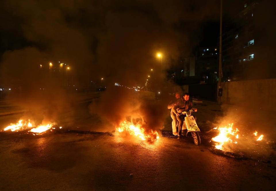 Lebanese citizens ride their scooter between burning tires that were set on fire by Sunni protesters, at one of the major roadways between Beirut and the capital's southern suburbs, a Hezbollah stronghold, to show their support with Sunni town of Arsal, in Beirut, Lebanon, Tuesday, March 18, 2014. Gunmen from Lebanon's militant Hezbollah group and local Shiite Muslim residents tightened their blockade of a Sunni town of Arsal near the Syrian border Tuesday, sparking concerns that thousands of Syrian refugees stranded in the area could be cut off from humanitarian aid. (AP Photo/Hussein Malla)