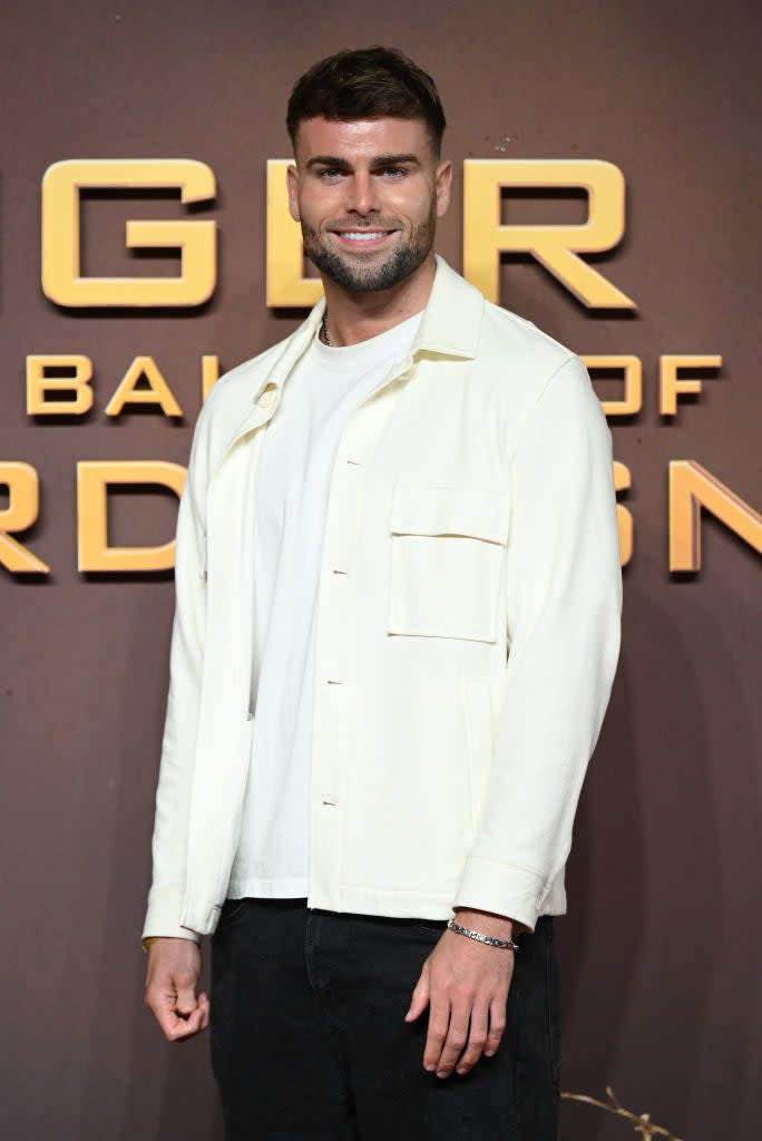 Tom in a casual light jacket over a tee with hands lightly clasped, standing before a promotional backdrop