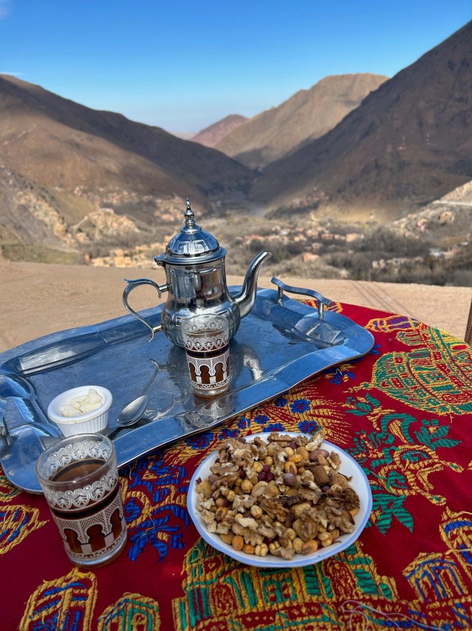 Traditional welcome fare in Morocco is shown here – mint tea and walnuts on a silver tray resting on a colorful tapestry. The drink is poured with ceremonial fanfare, holding the teapot spout high above the glasses.