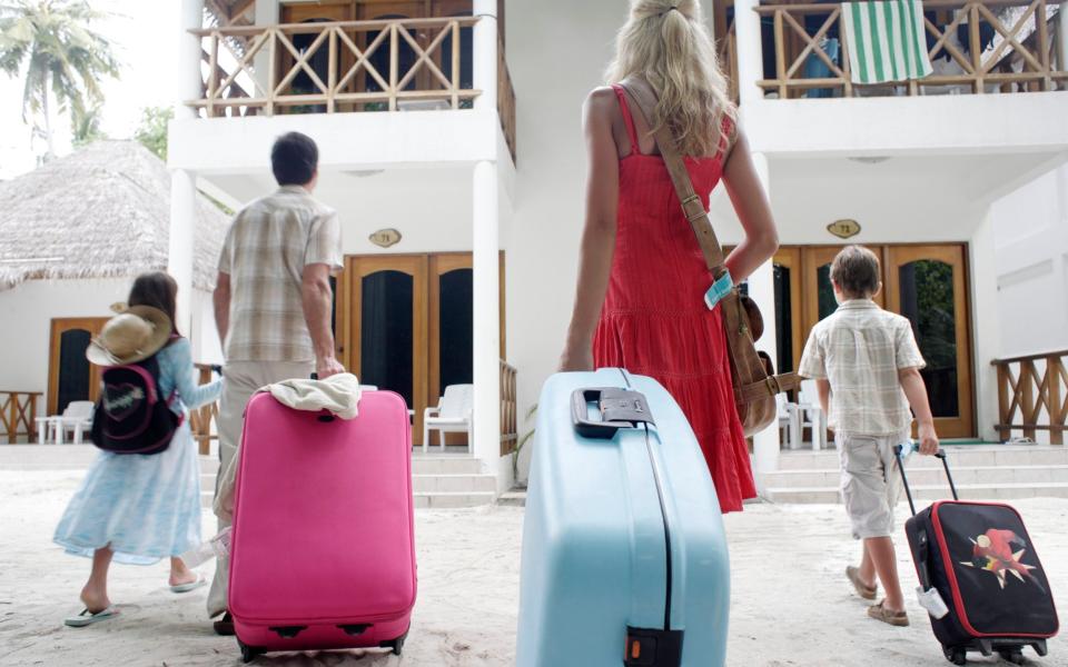 Family walking at resort with suitcase, rear view, how to save money on holidays - Getty