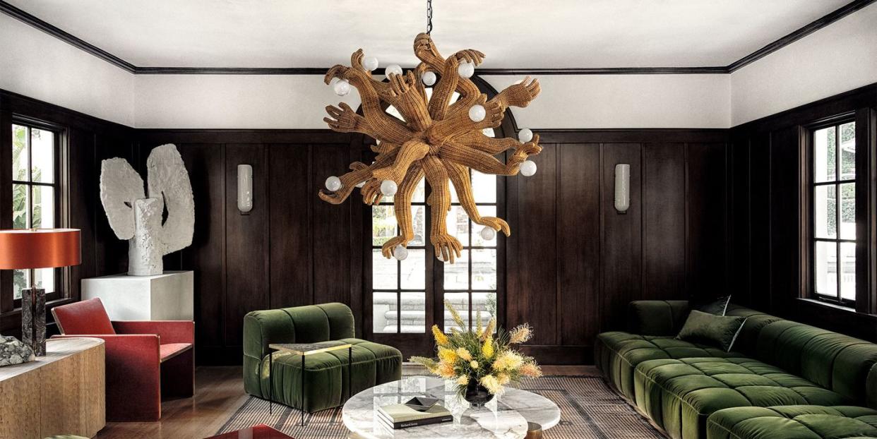 dark wood panelled living room with chandelier made of multiple arms and hands holding lights and a large green velvet tufted armless sofa and chairs and a low cocktail table at center with marble top and wooden legs