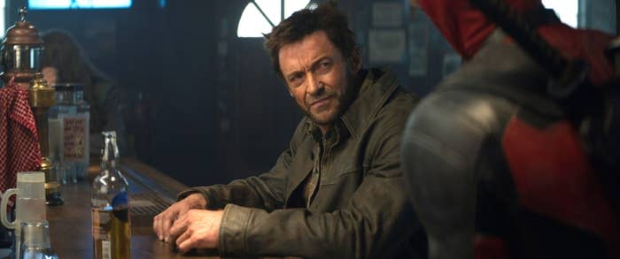 Hugh Jackman, wearing a casual jacket, sits at a bar counter and looks at Ryan Reynolds, dressed as Deadpool