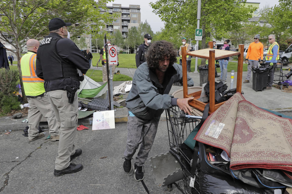 FILE - In this May 4, 2020 file photo a Seattle Police officer and other workers look on as a man wheels away a cart of his belongings from a homeless encampment being cleared, at Ballard Commons Park in Seattle. An affluent Seattle-area community has approved an ordinance prohibiting camping on public property. The city of Mercer Island says the measure will connect homeless people to shelters, but it might prove hard to enforce because of a federal court ruling. (AP Photo/Ted S. Warren,File)