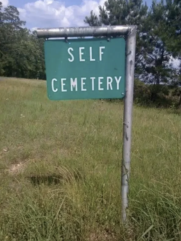 Sign reading "SELF CEMETERY" on a grassy area with trees in the background
