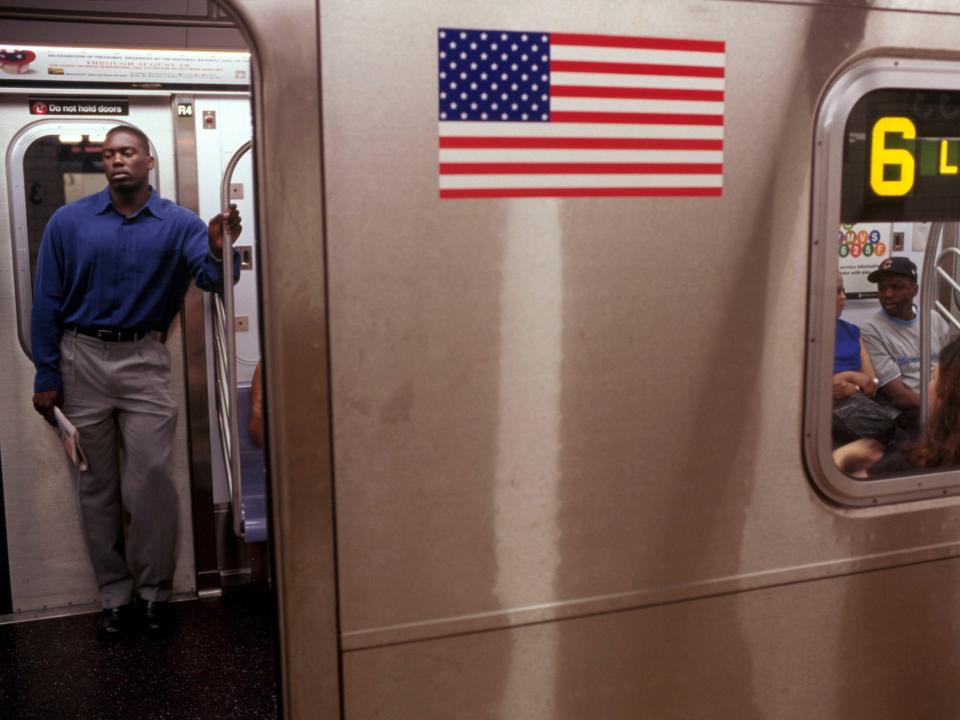 A man on the left stands inside a 6 train with the doors open and an American flag is seen to the right of the door on the exterior