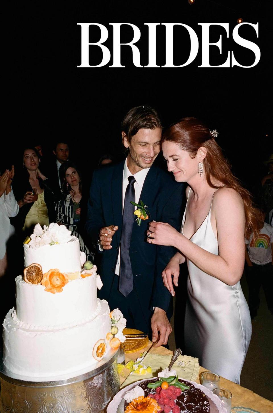 A bride and groom laugh as they stand next to their wedding cake.