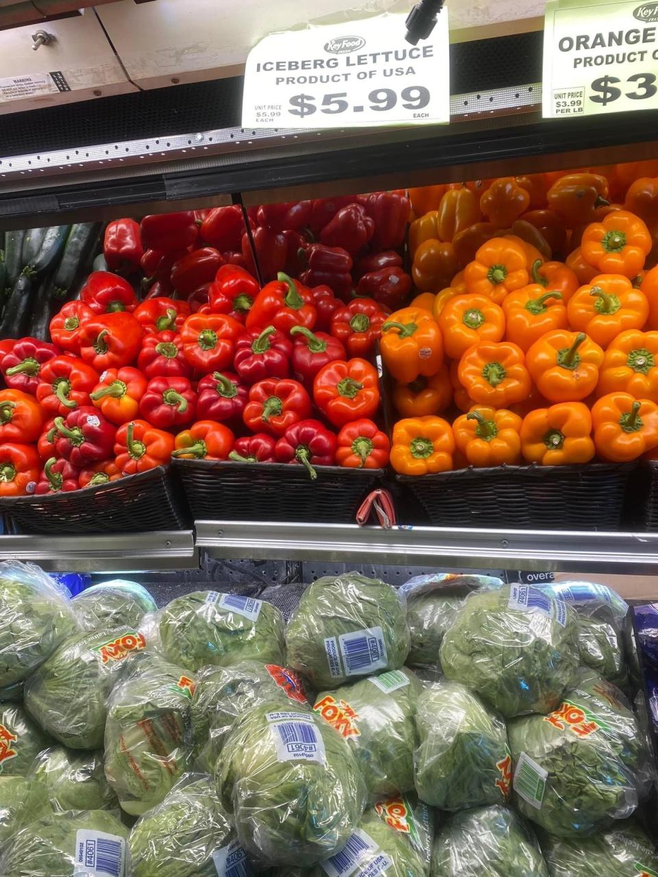 Grocery section displaying red and orange peppers on top shelves and packaged iceberg lettuce below, with price signs visible