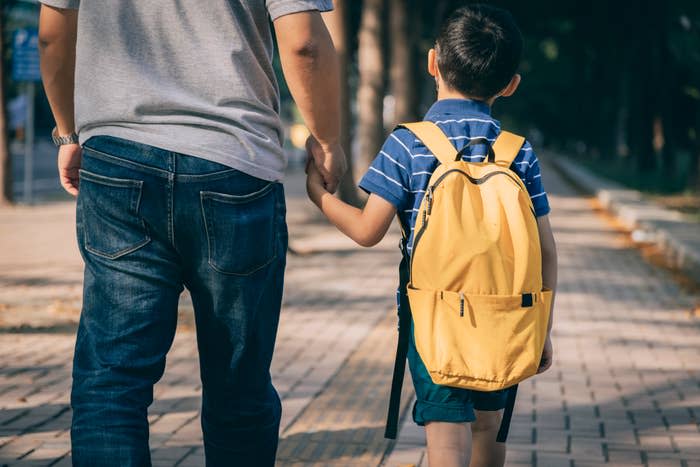 A man and a young boy with a yellow backpack walk hand-in-hand on a paved path. The boy wears a striped shirt. Their faces are not visible