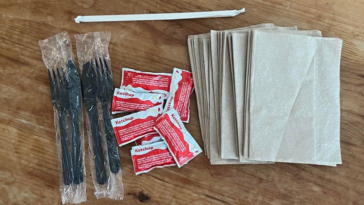 single-use take-out trash on table including napkins, ketchup packets, black plastic utensils in plastic and a straw