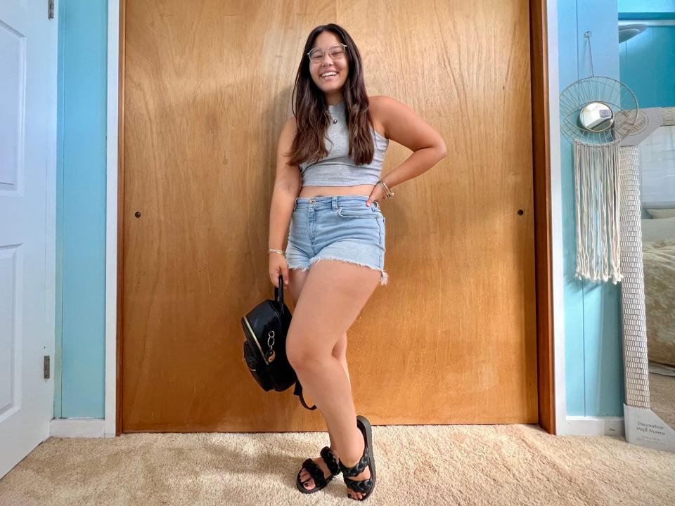 Author poses while wearing grey top, jean shorts, black backpack, and black sandals.