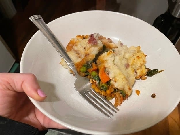 A person's hand holding a plate with a portion of shepherd's pie, including vegetables and mashed potatoes