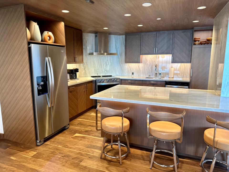 Stainless-steel fridge, wood cabinets with diagonal design and three stools next to marble countertop