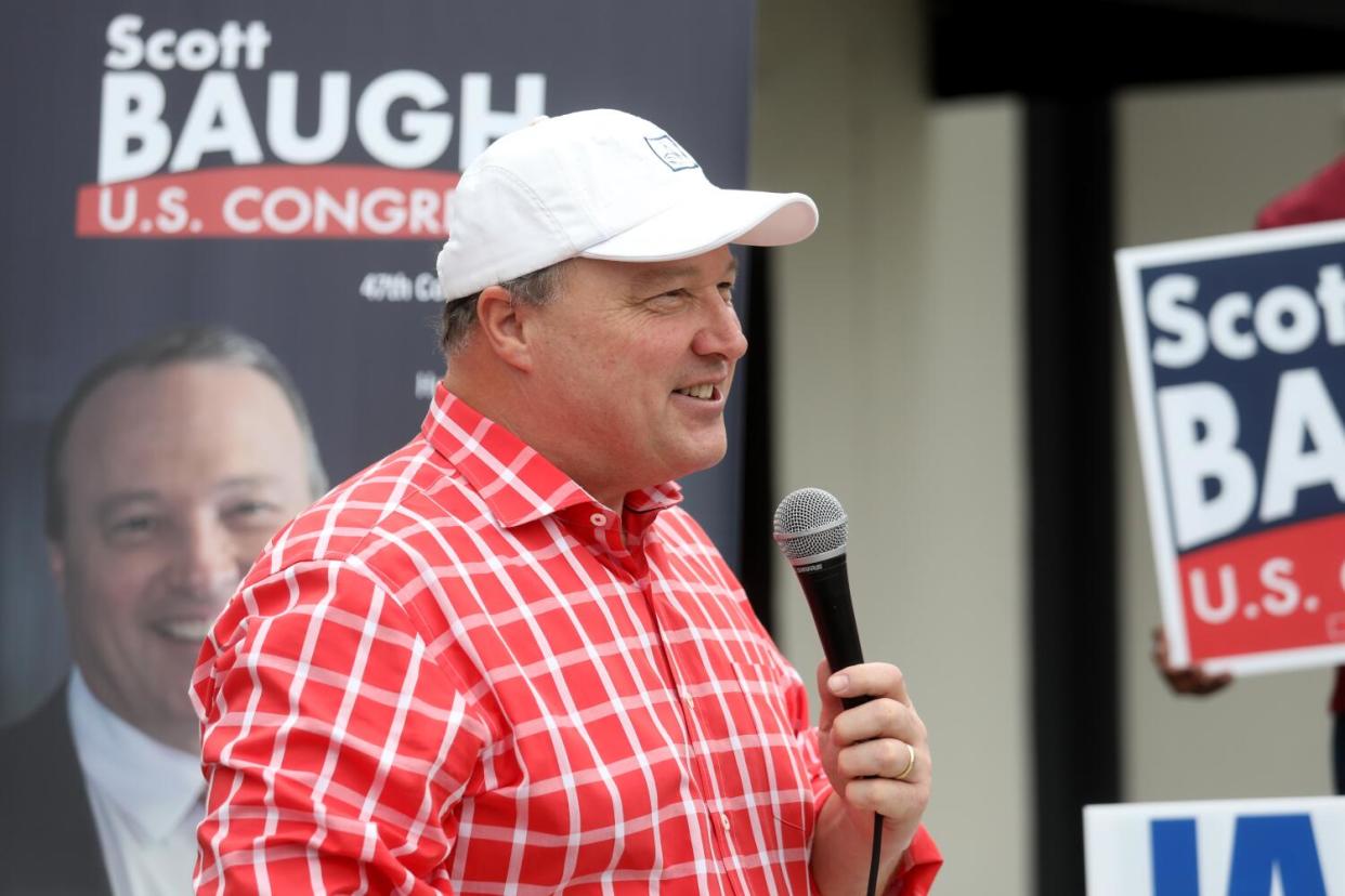Scott Baugh, in a red-and-white plaid shirt and white baseball cap, holding a mic as he speaks in front of campaign posters