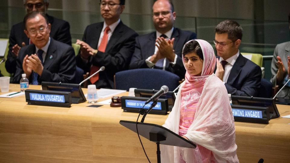 malala yousafzai stands at a podium with a microphone and looks outward as men sitting behind her clap, she wears a pink outfit