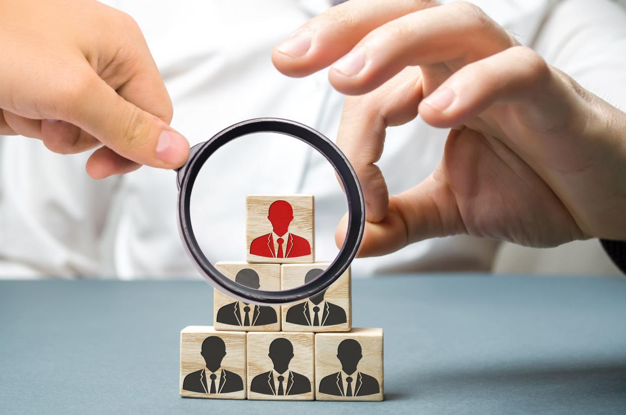 A magnifying glass is held in front of the top wooden block in a pyramid of wooden blocks with businessmen on them.