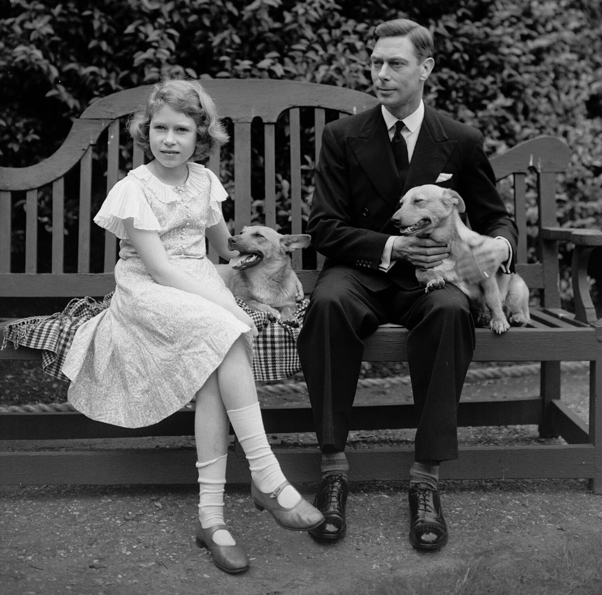 July 1936: George, Duke of York and Princess Elizabeth sitting on a bench with their corgi dogs in the grounds of their London home, 145 Piccadilly.