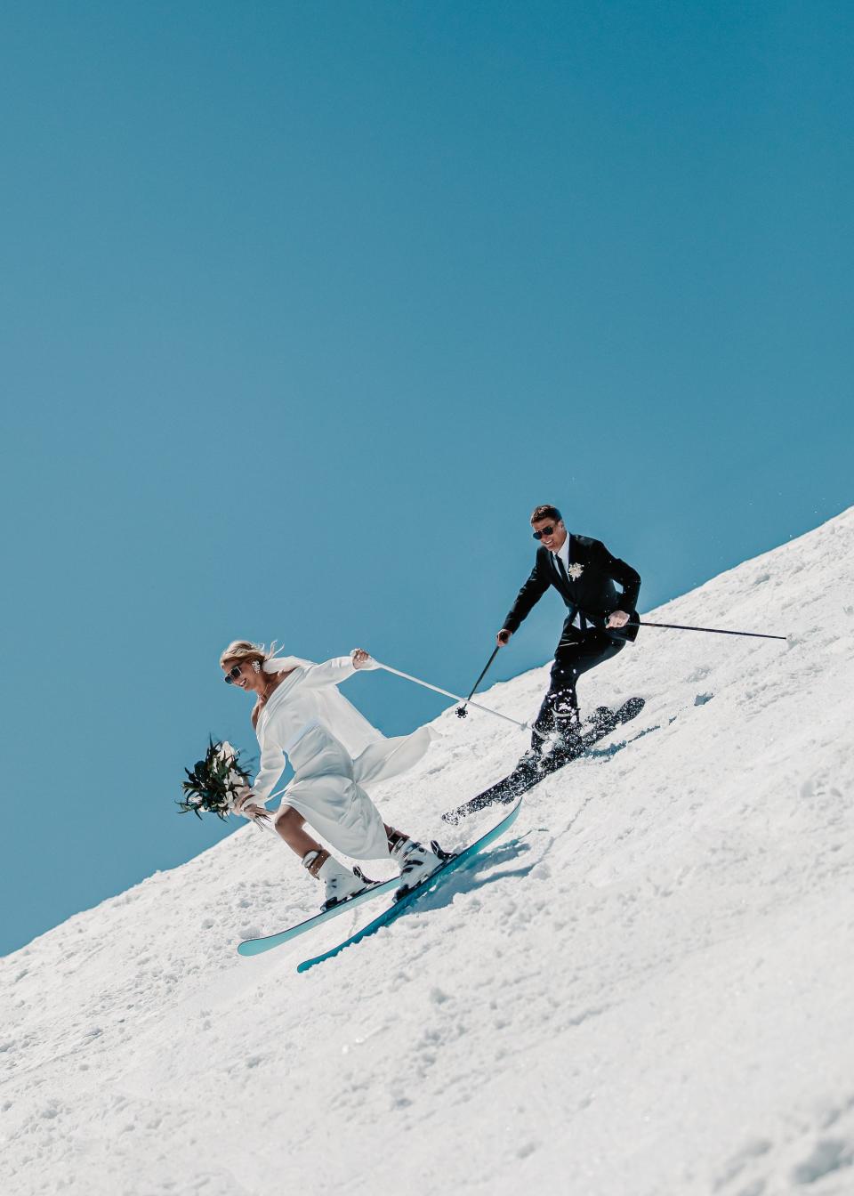 A bride and groom ski down a mountain in their wedding attire.