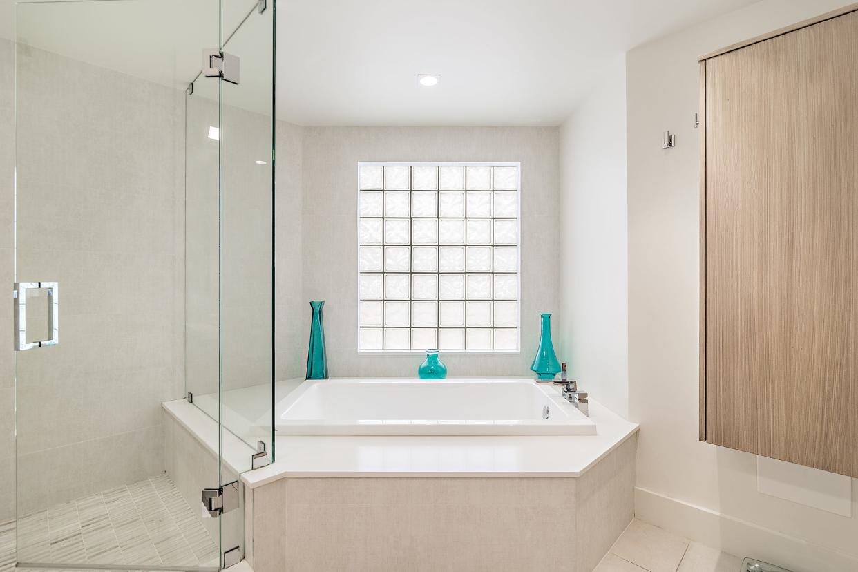 In the primary bathroom, the bathtub is positioned beneath a glass-block window and next to the glass-enclosed shower.