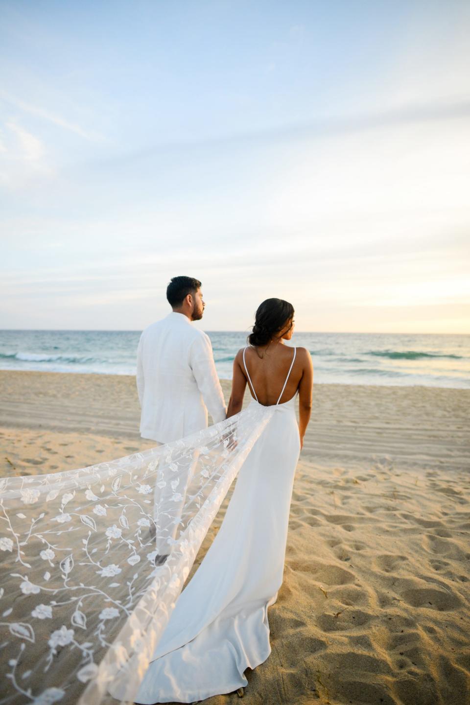 A man in a white suit and a woman in a wedding dress look at the ocean while standing on the beach.
