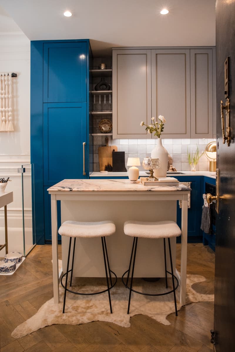 Kitchen in new york apartment after renovation/decorating. Blue tall cabinets, gray cabinets over sink, white marble island with 2 stools, shelves for glasses, etc. White countertops and tile backsplash. Brass accents and hardware