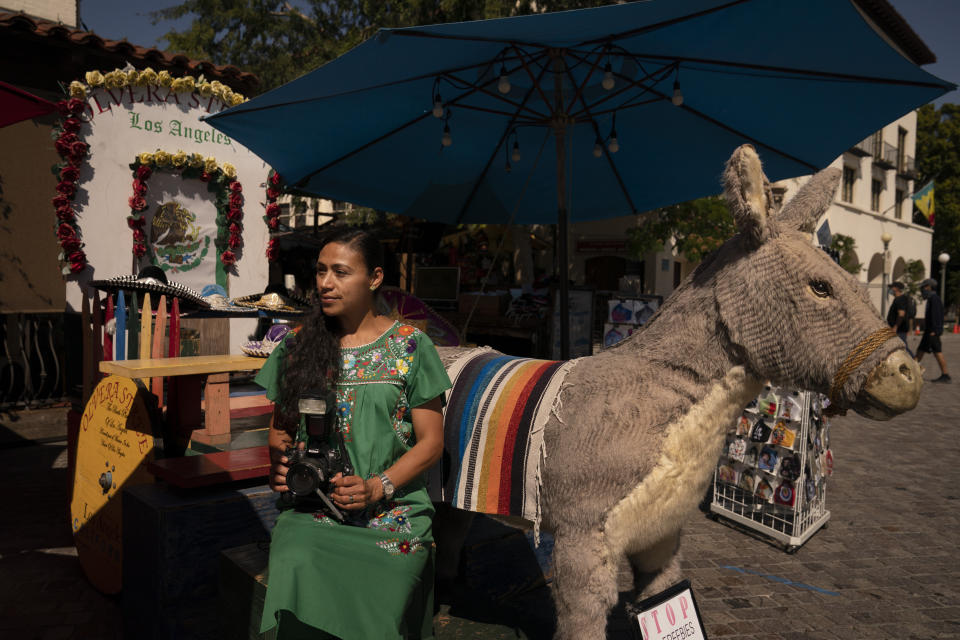 Photographer Carolina Hernandez, 39, sits for a photo next to a stuffed donkey, a photo prop named George, at her kiosk on Olvera Street in Los Angeles, Friday, June 4, 2021. As Latinos in California have experienced disproportionately worse outcomes from COVID-19, so too has Olvera Street. The shops lining the narrow brick walkway rely heavily on participants at monthly cultural celebrations, downtown office workers dining out, school field trips and Dodger baseball fans loading up on Mexican food before or after games. But the coronavirus killed tourism, kept office workers and pupils at home, cancelled events and kept fans from sporting events. (AP Photo/Jae C. Hong)