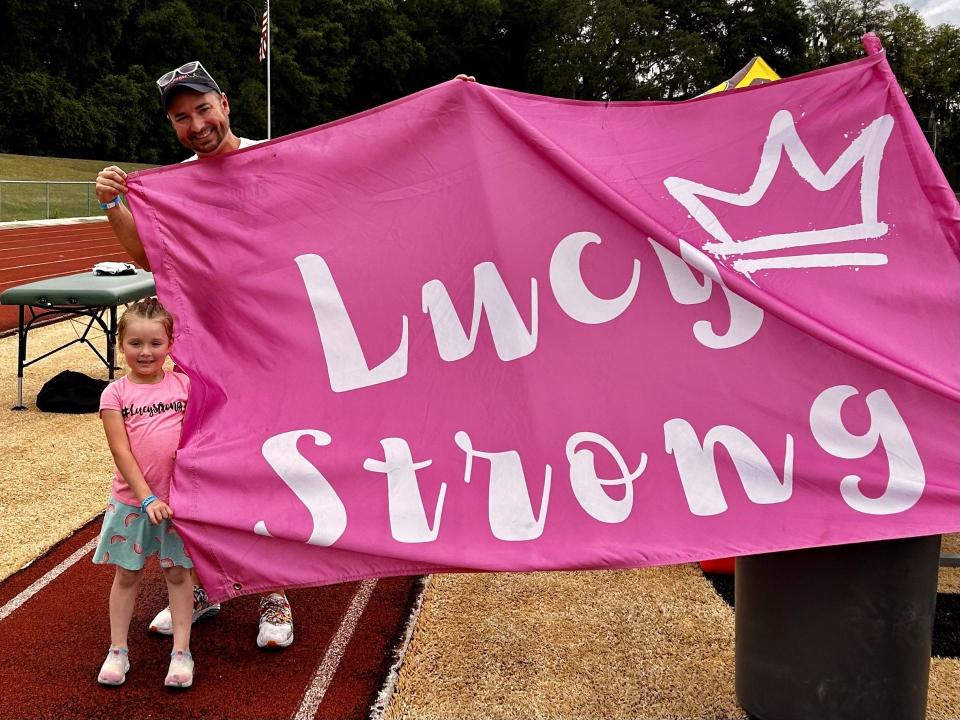 Jordan Donmoyer and his 5-year-old daughter Charlotte display the flag the Jacksonville University women's lacrosse team carries onto the field before every game as a reminder of Charlotte's sister Lucy, who died in September of 2023. The family connected with JU through Friends of Jaclyn, a program that pairs college teams with pediatric cancer victims.