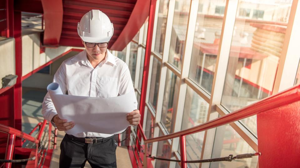 Engineer or Architect checking architectural drawing while wearing a personal protective equipment safety helmet at construction site.