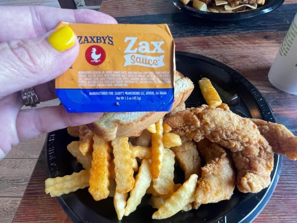 The writer holds a package of Zax sauce with a plate of fries and chicken fingers in the background