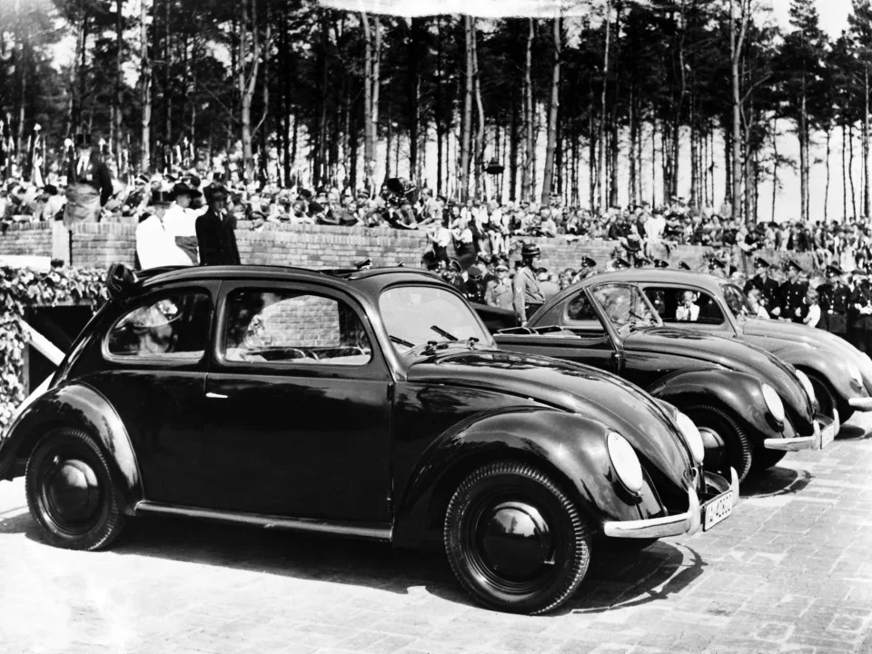 Adolf Hitler stands behind the cars in a dark coat, preparing to dedicate the new factory.