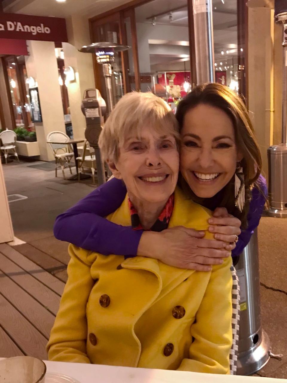 Barbara Rush in a yellow jacket being hugged from behind by her daughter as they are both smiling