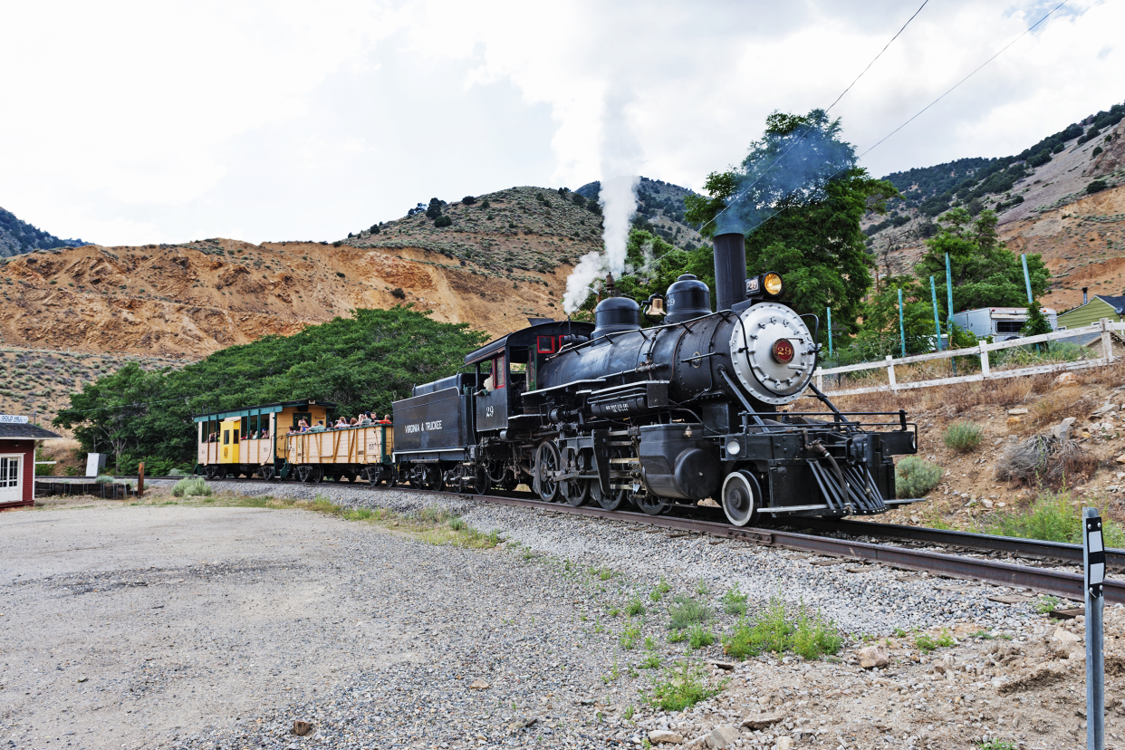 A train in motion with passengers on the railroad in Virginia City, Nevada, gravel in the foreground, mountains in the background, white clouds in the blue sky