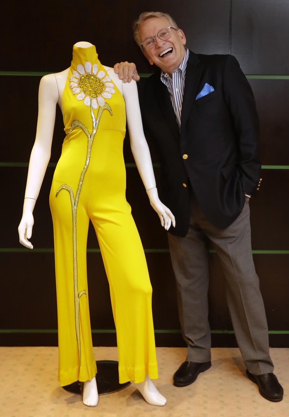 Fashion and Costume Designer Bob Mackie poses with one of his iconic designs, a marigold jersey jumpsuit worn by Cher between 1971-1976, in London, Thursday, Aug. 16, 2018. The jumpsuit which is embellished with a large white sunflower composed of sequins, and beads, is estimated at 3,000-5,000 US Dollars (2,363-3,938 UK Pounds) and will be auctioned in the 'Property from the Collection of Bob Mackie' sale by Julien's Auctions in Los Angeles on Nov. 17. (AP Photo/Kirsty Wigglesworth)