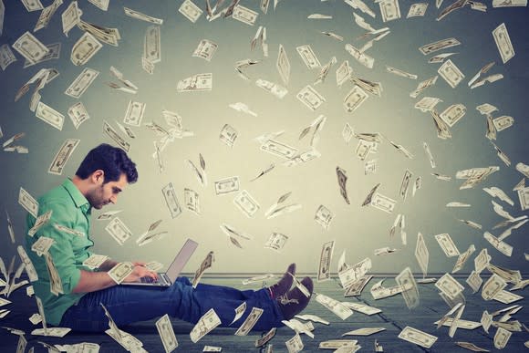 A man sits with his laptop as paper money falls around him