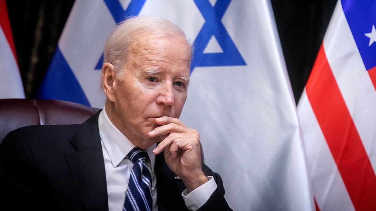 Biden with hand up to lips sitting in front of Israel flag