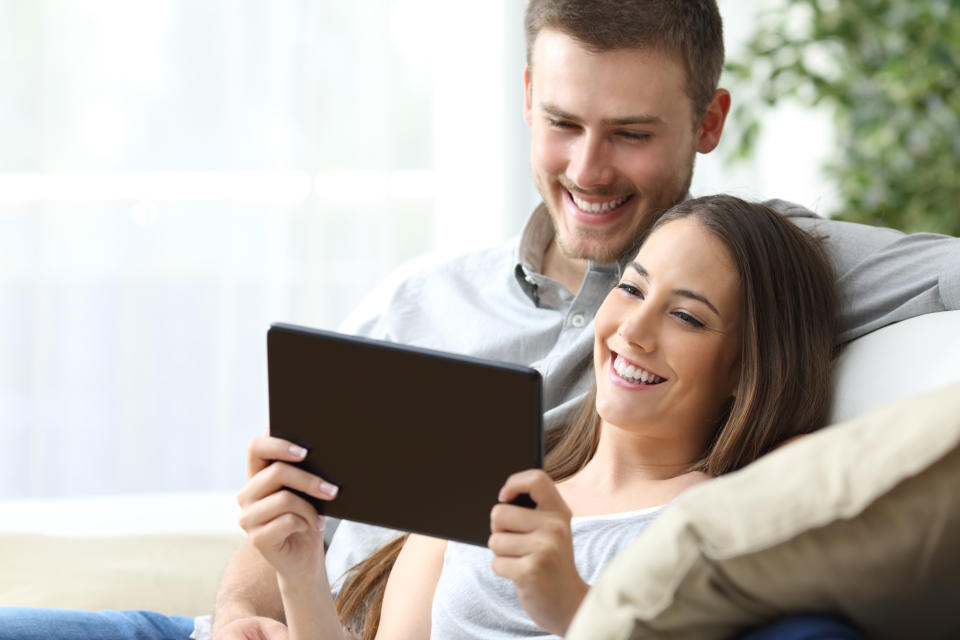 A young man and woman smiling while sitting on a couch looking at a tablet.