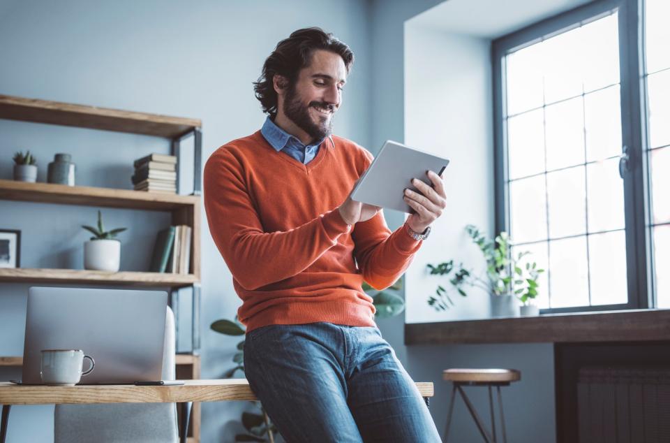 An investor leans against a desk at home and looks at something on a tablet.