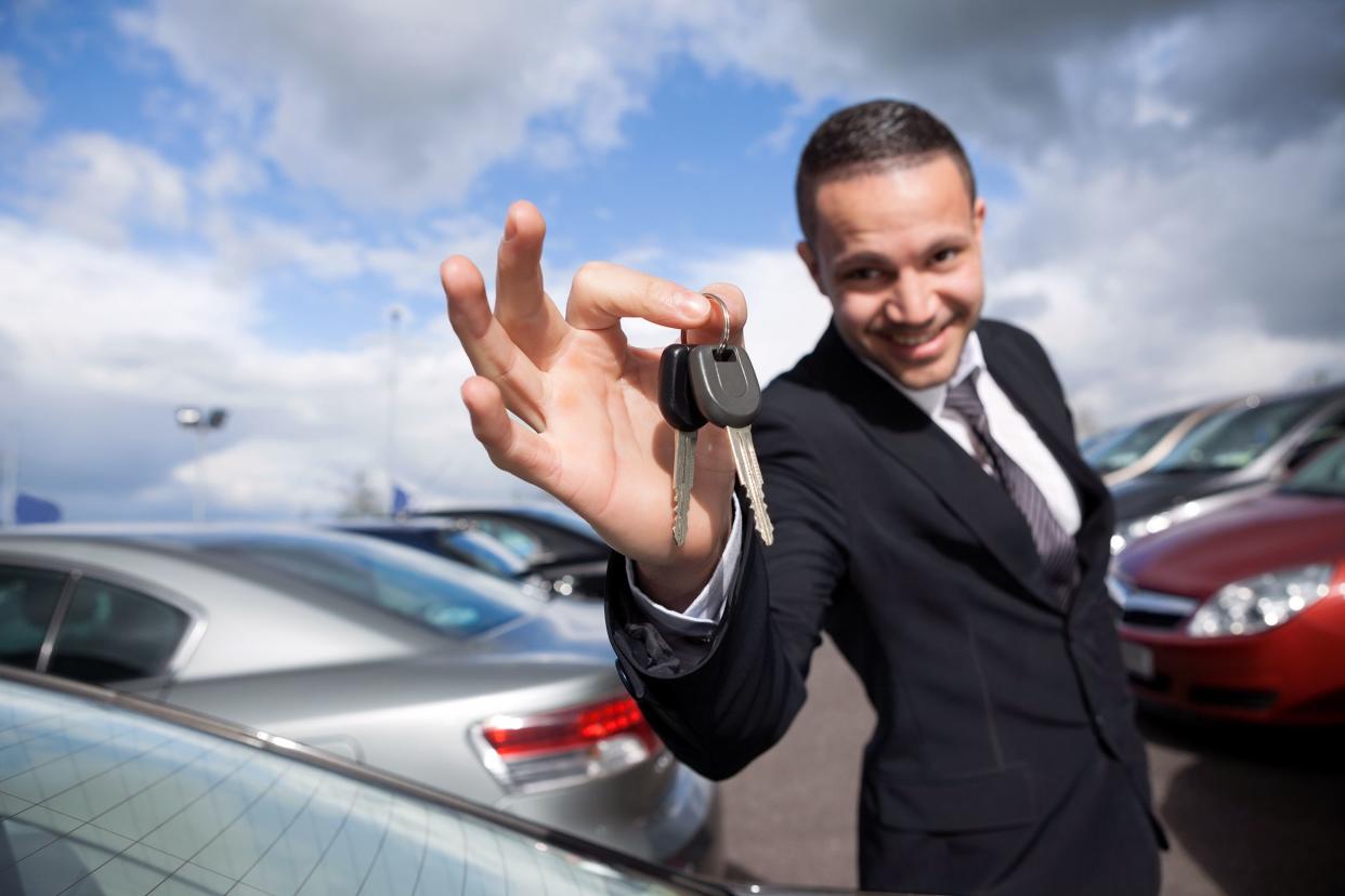 Happy male car dealer looking into camera while holding car keys with cars at car dealership in background