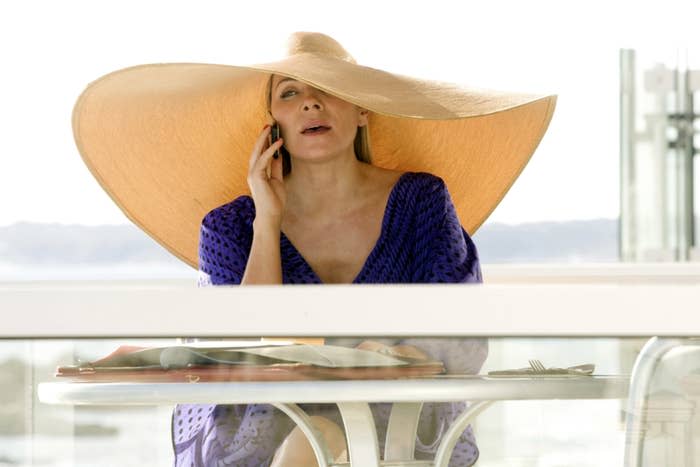 Samantha sitting at a balcony table and wearing an oversized sunhat while talking on the phone
