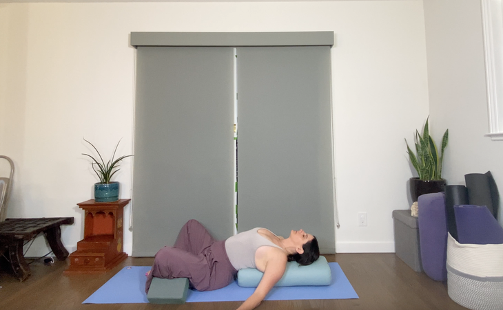 A woman in loose purple pants reclines on a bolster in a variation on Supta Baddha Konasana, Recliing Bound Angle Pose